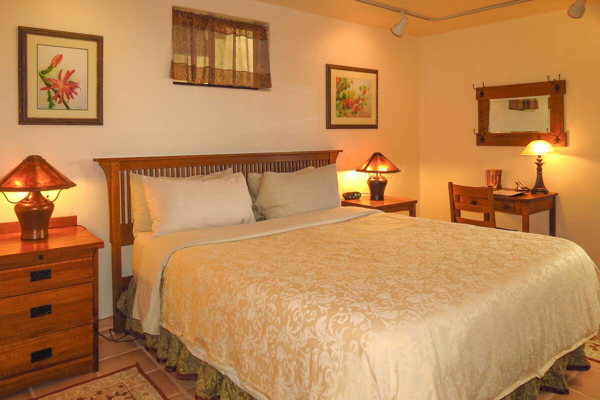A neatly arranged bedroom with a large bed, two bedside tables with lamps, a wooden nightstand, a chair, and a desk with a mirror.