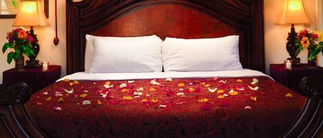A large bed with a dark wooden headboard, white pillows, and a red patterned bedspread. There are two bedside tables with lamps and flower arrangements.