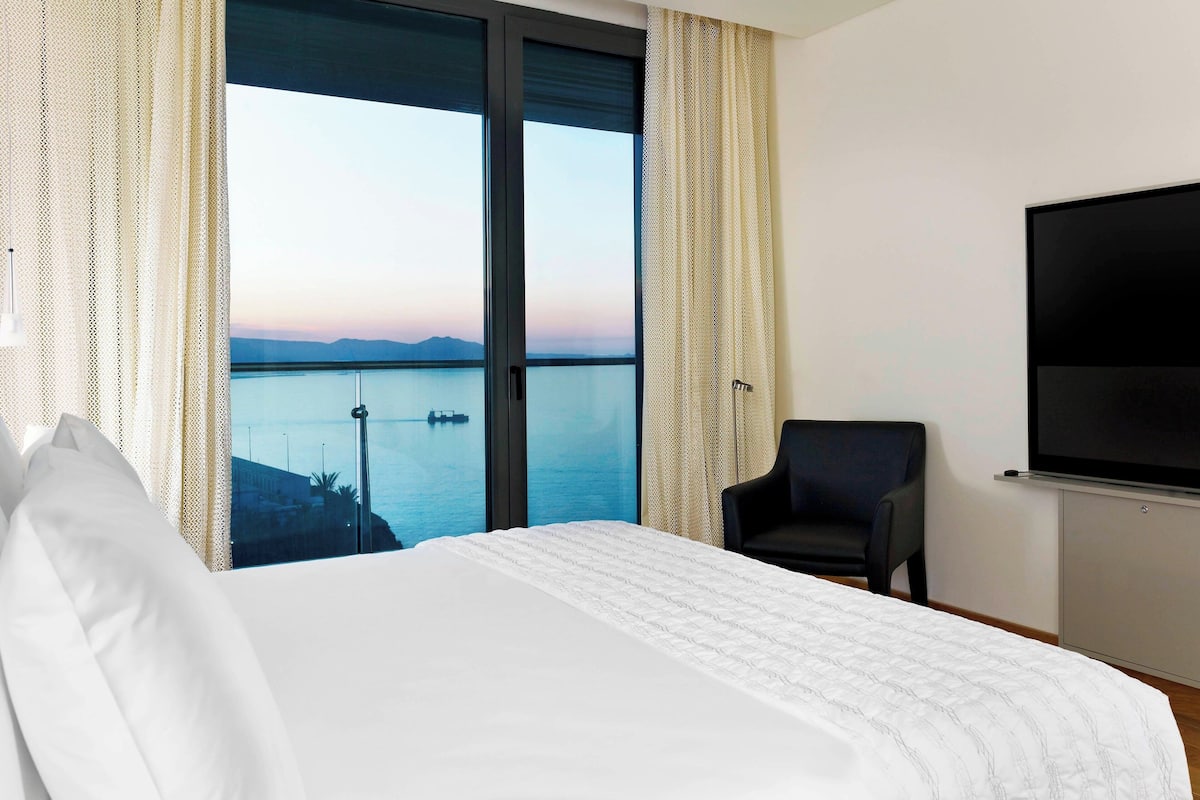 Une chambre d’hôtel avec un grand lit, une chaise, une télévision et une vue sur la mer.