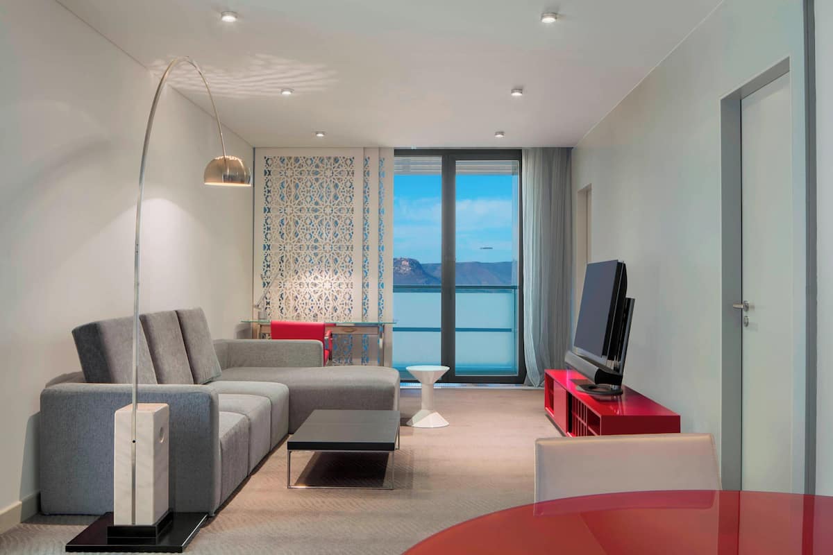 Un salon moderne avec un canapé d’angle, un téléviseur à écran plat posé sur un meuble rouge, une lampe sur pied et une vue sur les montagnes grâce à de grandes fenêtres.