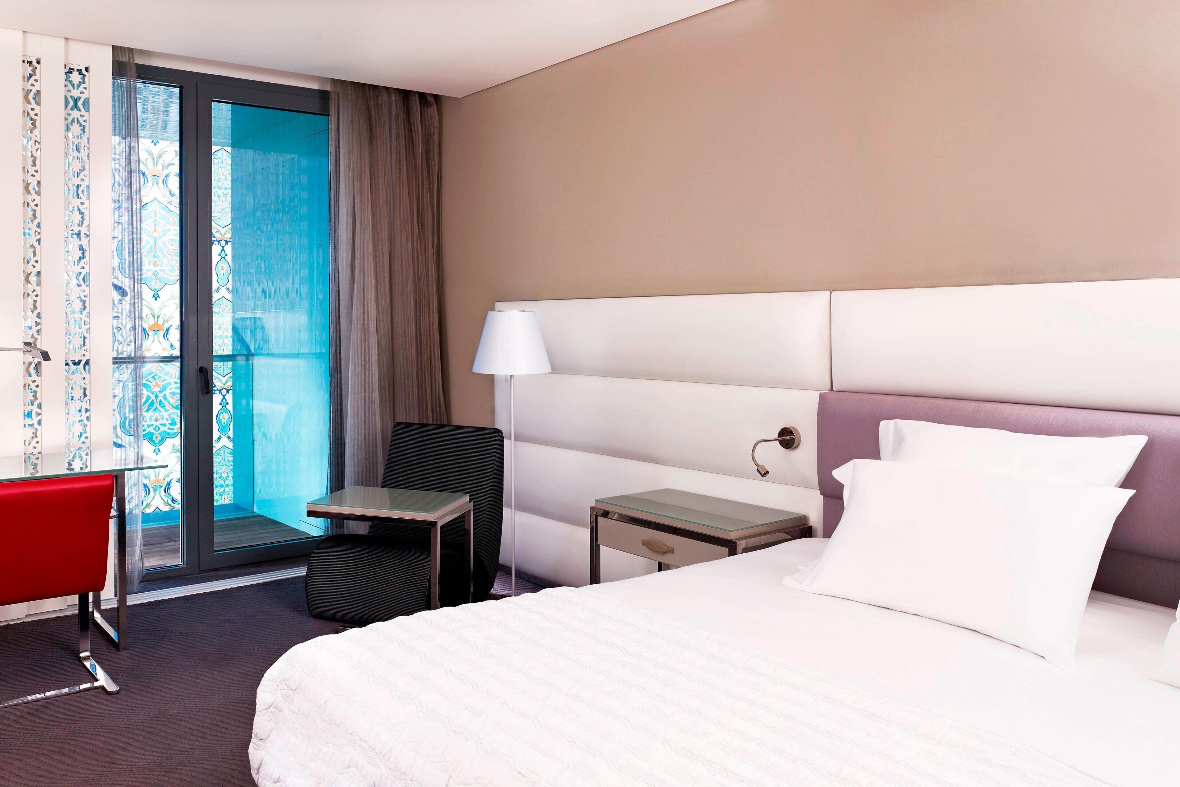 A hotel room with a large bed, a desk, a chair, and a view of the outdoors through a glass door.