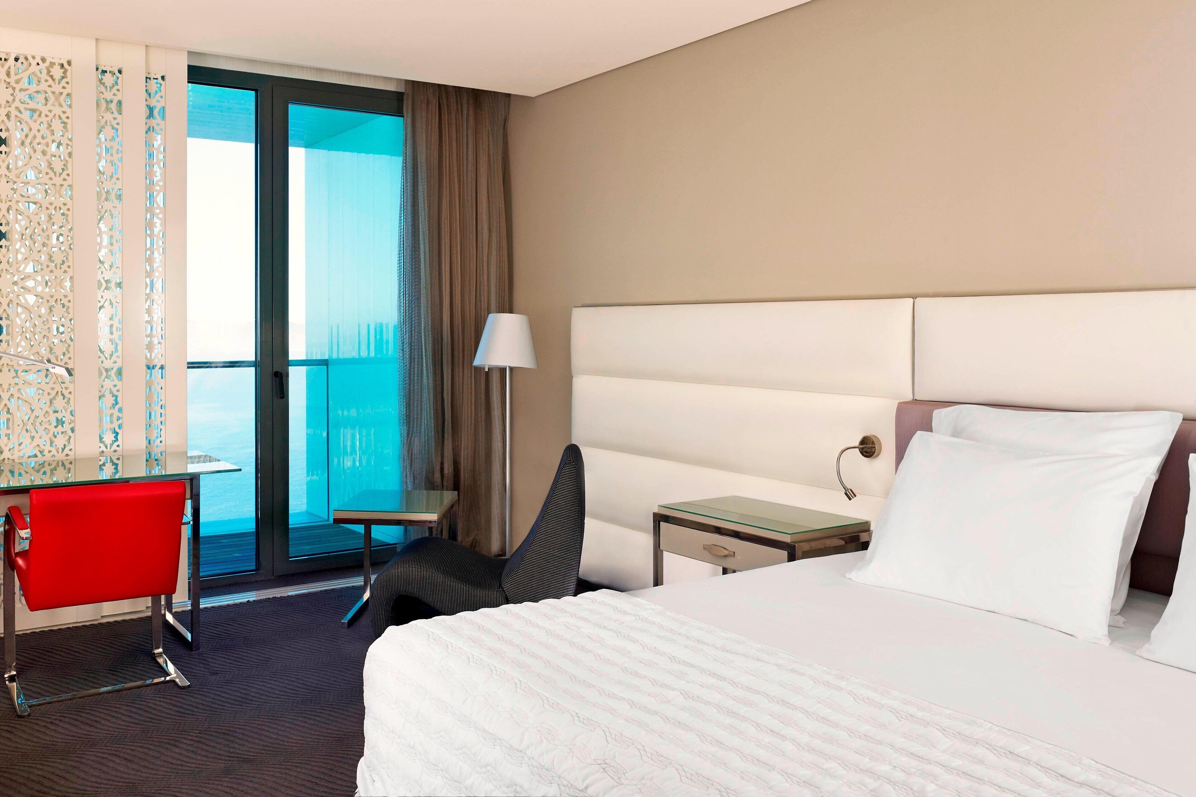 Une chambre d’hôtel avec un grand lit, un bureau, une chaise, une table de chevet et une vue sur l’océan par une porte vitrée.