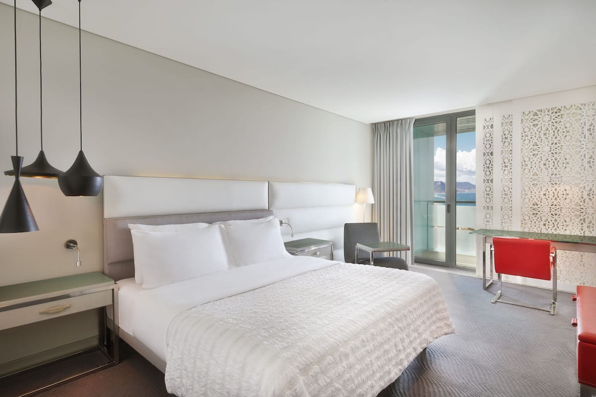 Une chambre d’hôtel avec un lit, une table de chevet, une lampe, une chaise, un bureau et une vue par la fenêtre.