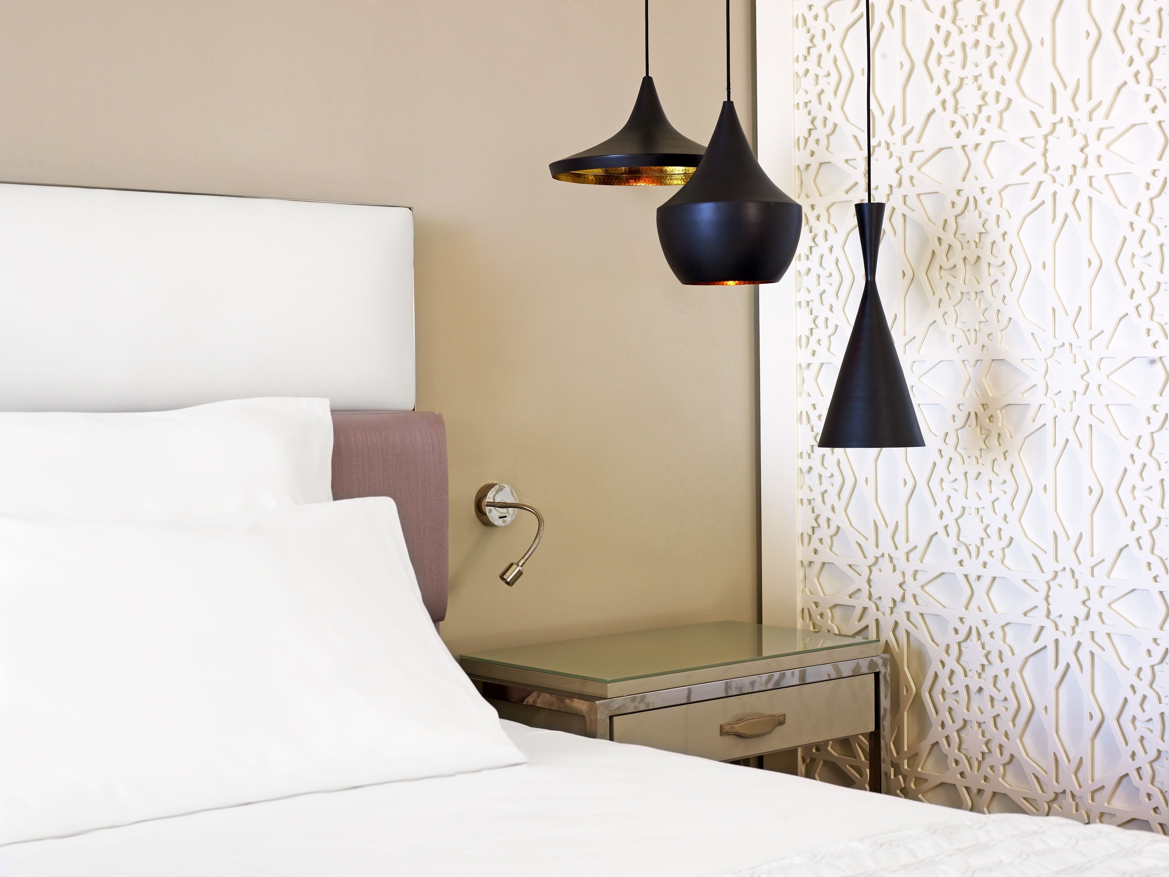 A neatly made bed with white and pink linens, a bedside table with a glass top, and two pendant lights hanging above.
