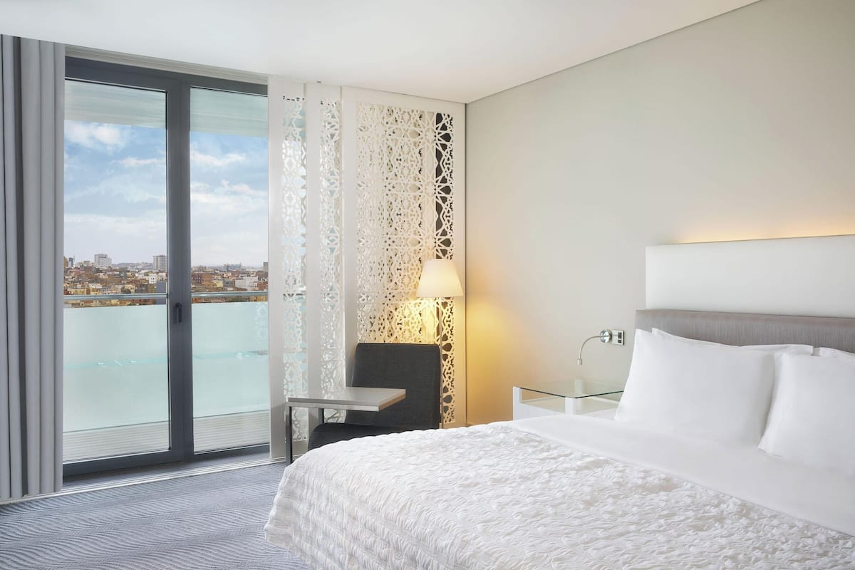 Une chambre d’hôtel avec un grand lit, une chaise, une petite table, une lampe et une vue sur la ville par une grande fenêtre.