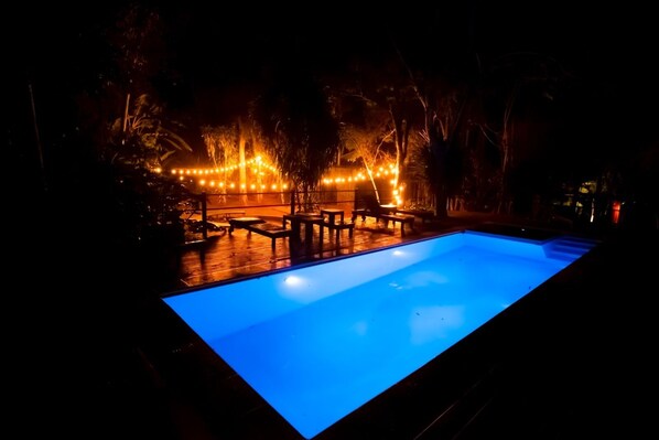 Una piscina con muelle de madera y sillas de descanso, rodeada de luces colgantes.