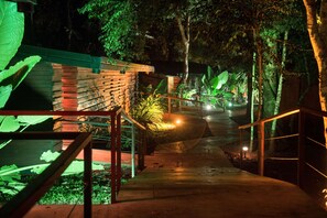 Un sendero nocturno con muro de ladrillo, barandilla metálica y plantas iluminadas.