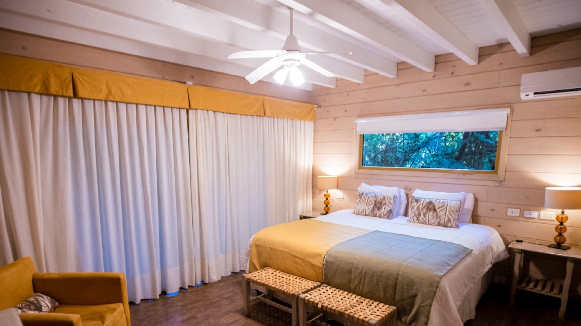 A bedroom with a bed, a bench, a chair, a window with a view, and a ceiling fan.