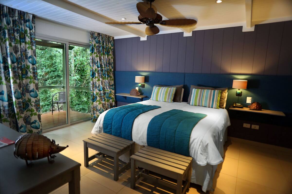A bedroom with a large bed, two bedside tables with lamps, a ceiling fan, and a view of greenery through the glass door.