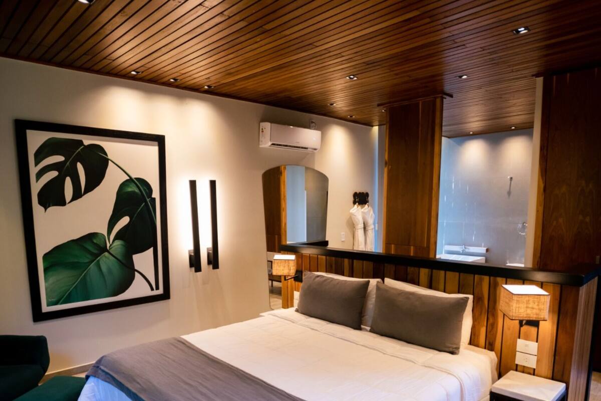 A hotel room with a bed, a large plant artwork, a mirror, and a wooden ceiling.