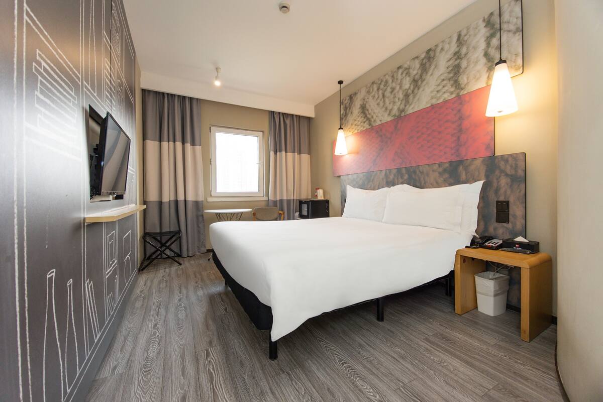 A hotel room with a bed, bedside table, lamp, TV, and a window with curtains.