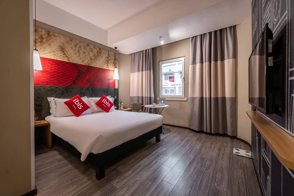 Ein Hotelzimmer mit Bett, Schreibtisch, Stuhl, Fernseher und einem Fenster mit Vorhängen.