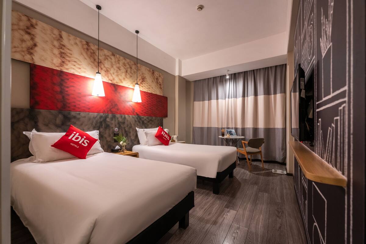 Ein Hotelzimmer mit zwei Betten, einem kleinen Schreibtisch, einem Stuhl und einem an der Wand befestigten Fernseher.