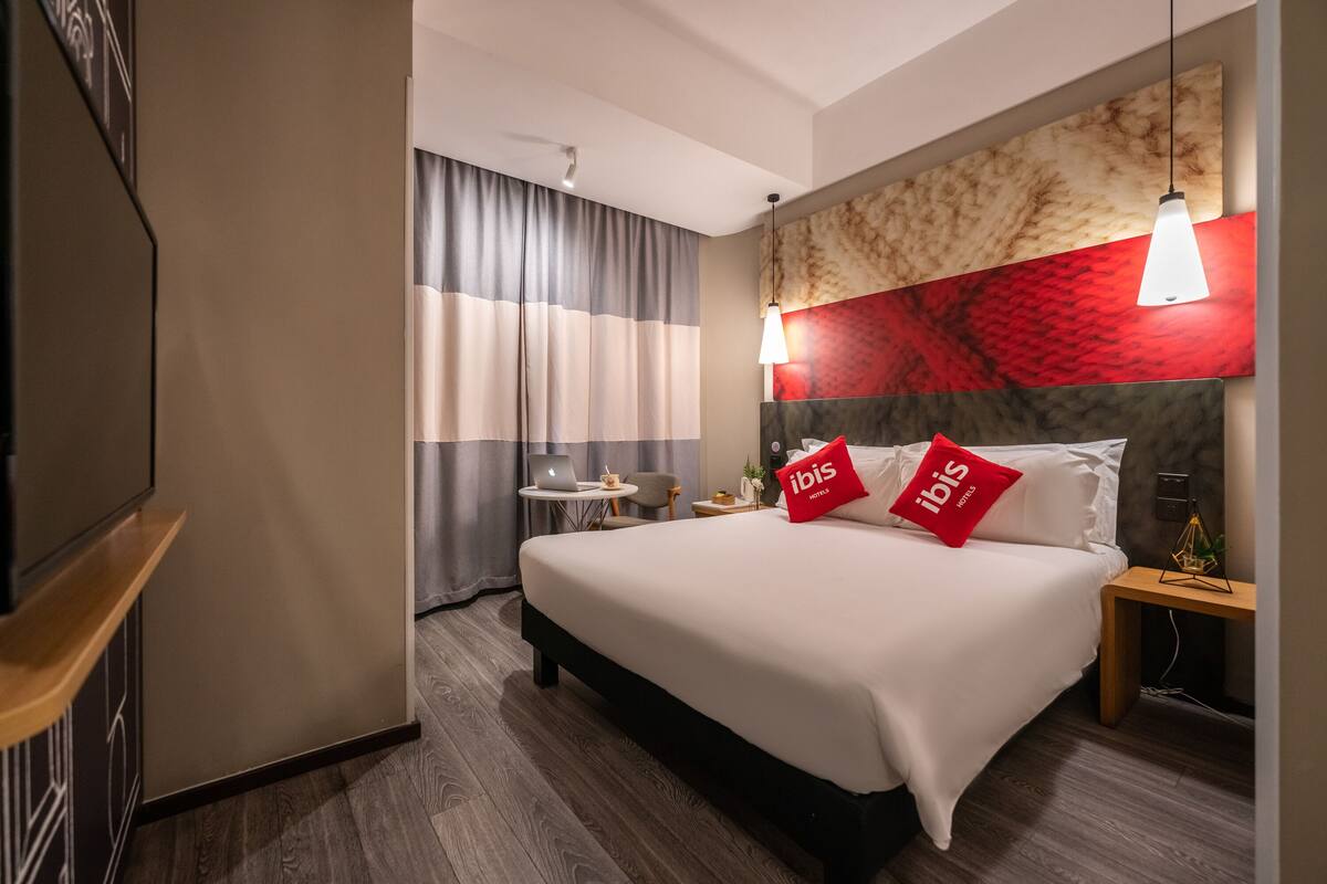 Ein Hotelzimmer mit einem großen Bett, einem Schreibtisch mit einem Laptop, einem kleinen Tisch mit einer Lampe und einer Wand mit einem rot-beigen Muster.
