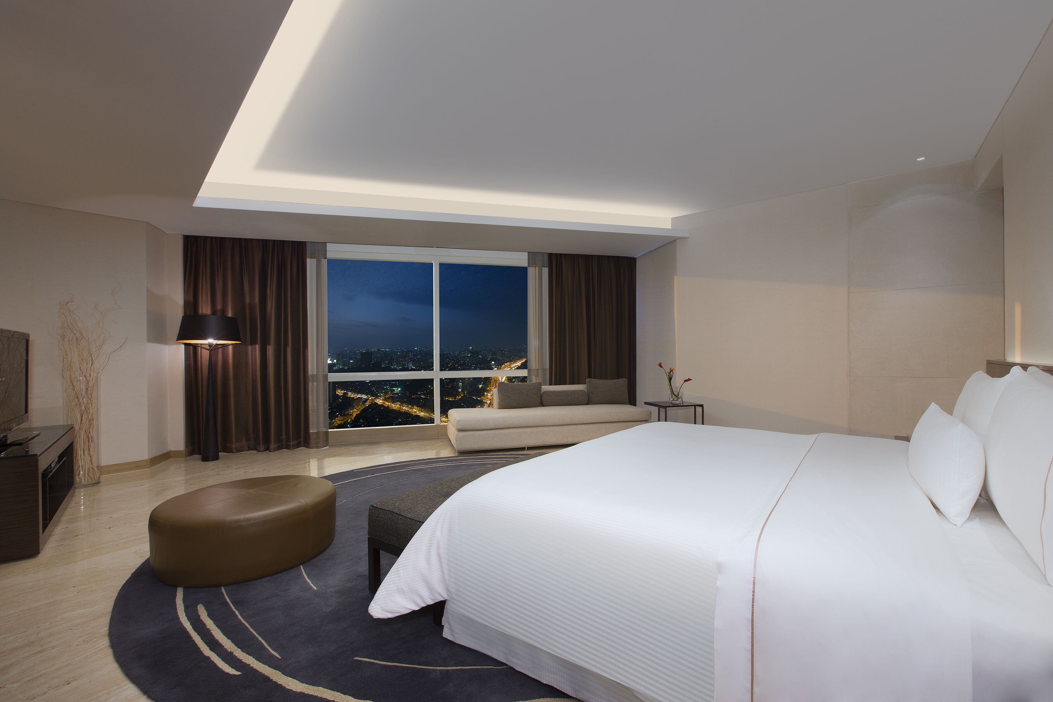 A modern hotel room with a large bed, a seating area, a desk, and a view of the city at night.