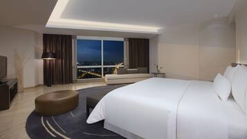 A modern hotel room with a large bed, a seating area, a desk, and a view of the city at night.