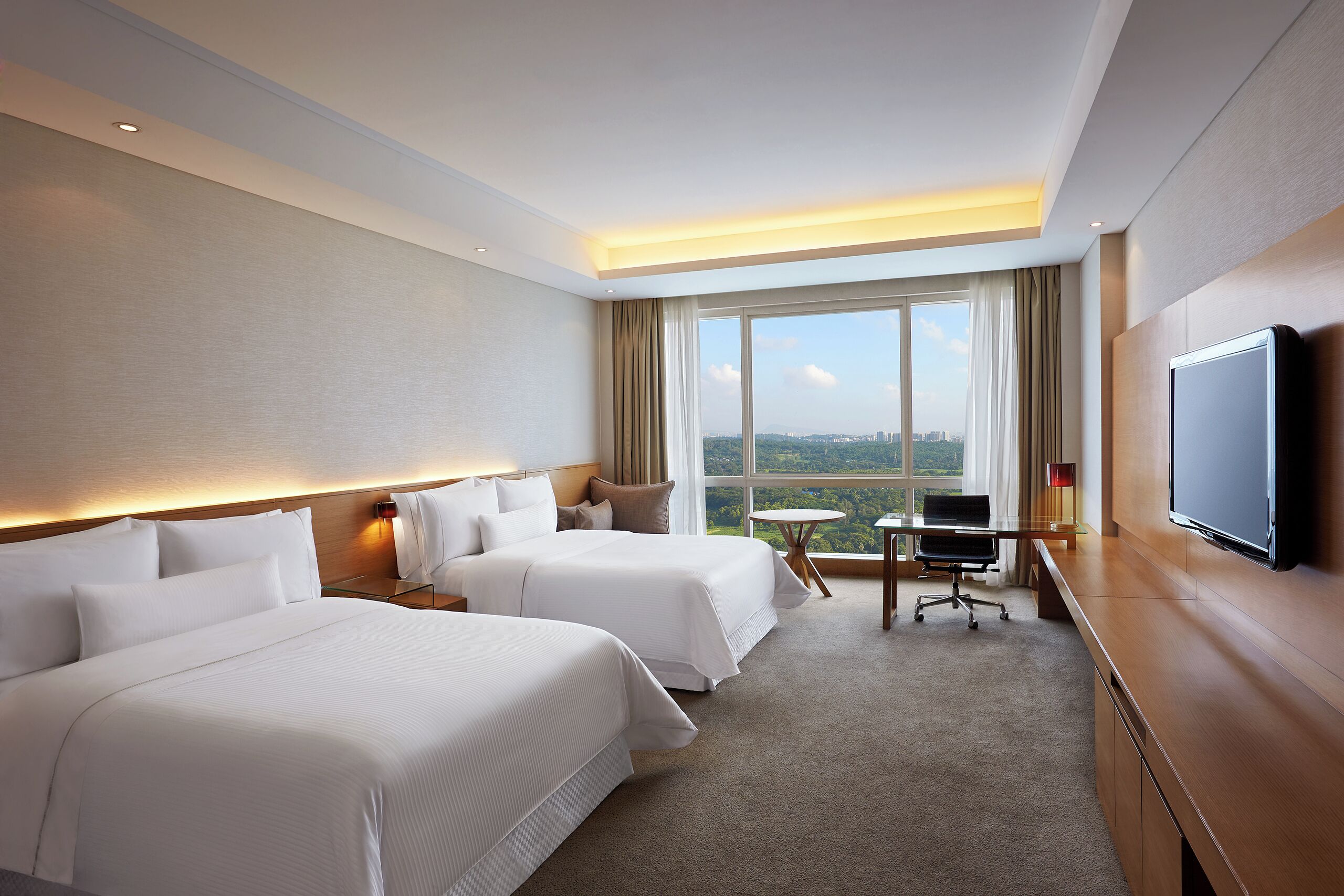 A hotel room with two beds, a desk, a chair, a TV, and a large window offering a city view.