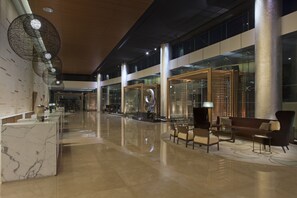 Un vestíbulo amplio con una recepción de mármol, zona de estar con sofás y sillones, y grandes puertas de cristal que dan a otras áreas.