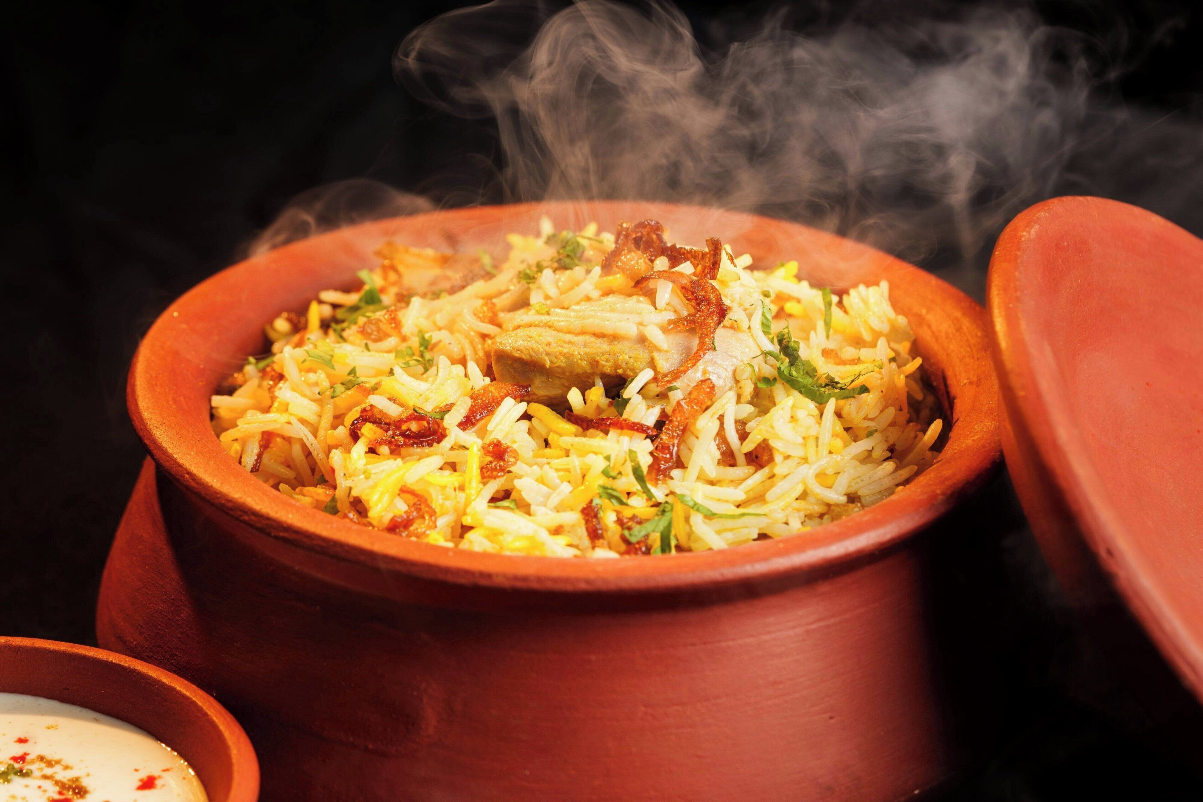 A clay pot filled with biryani, steam rising from the dish, and a small bowl of yogurt on the side.