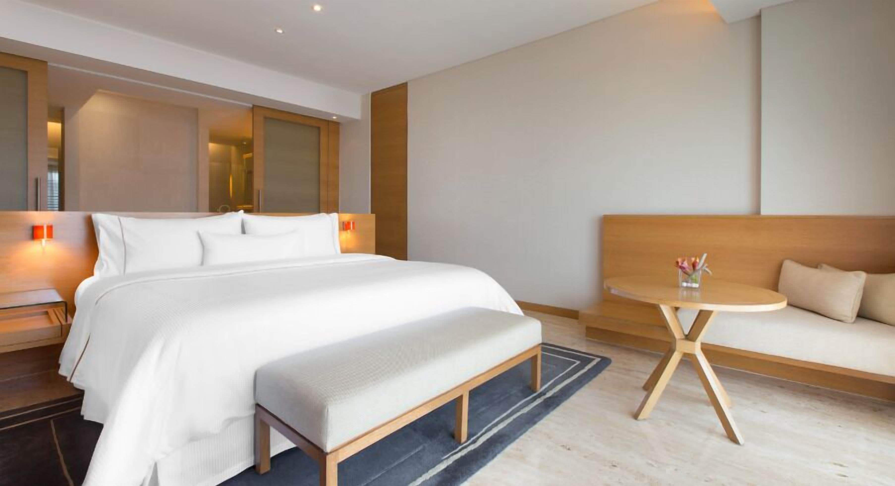 A modern hotel room with a large bed, a sofa, a small round table, and a bench.