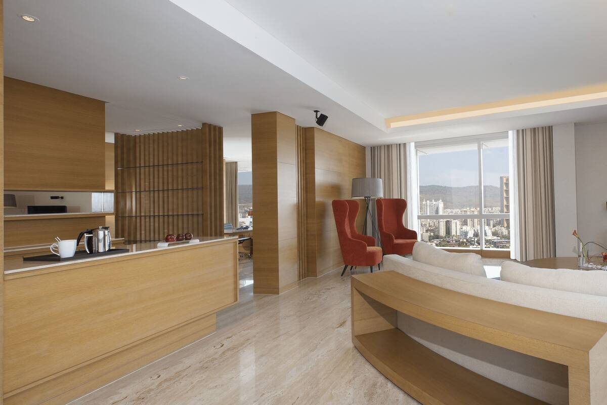 A modern hotel room with a kitchen area, a seating area with a sofa and chairs, a TV, and a large window offering a city view.