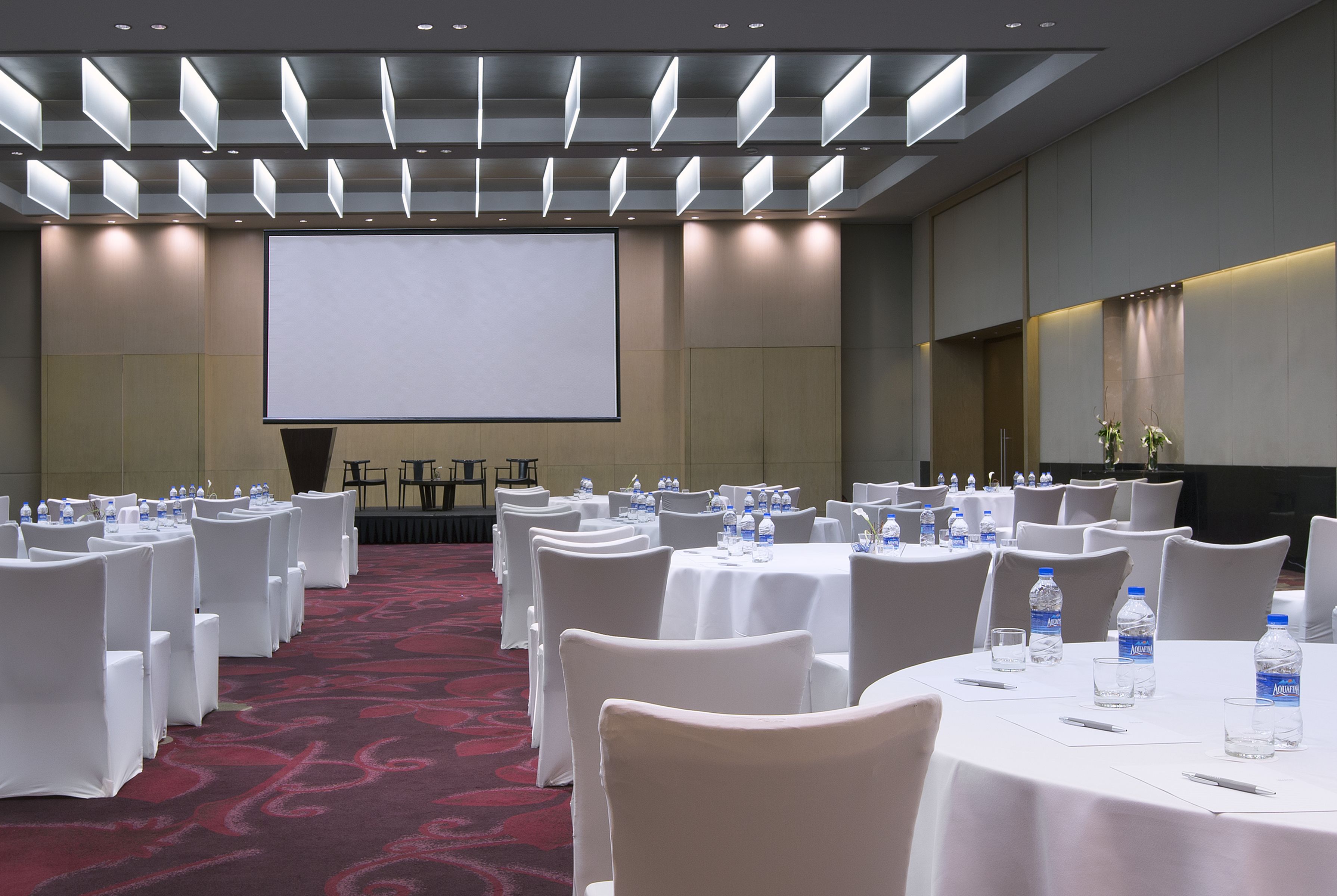 Un salón de conferencias con filas de mesas y sillas blancas, una pantalla grande y botellas de agua en cada mesa.