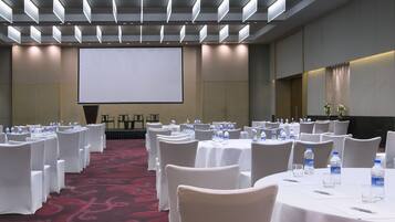 Un salón de conferencias con filas de mesas y sillas blancas, una pantalla grande y botellas de agua en cada mesa.