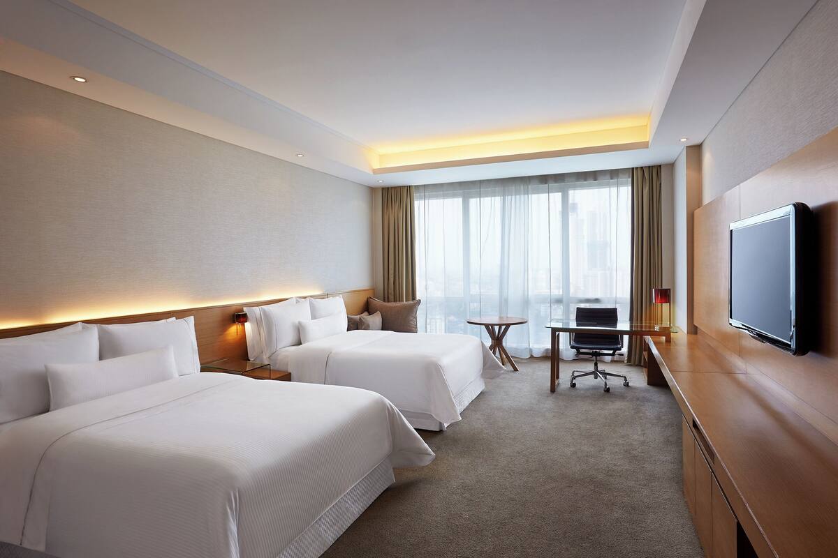 A hotel room with two beds, a desk, a chair, a TV, and a large window with curtains.
