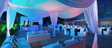 An outdoor event space with draped fabric ceilings, white cushioned seating, and tables set for dining.