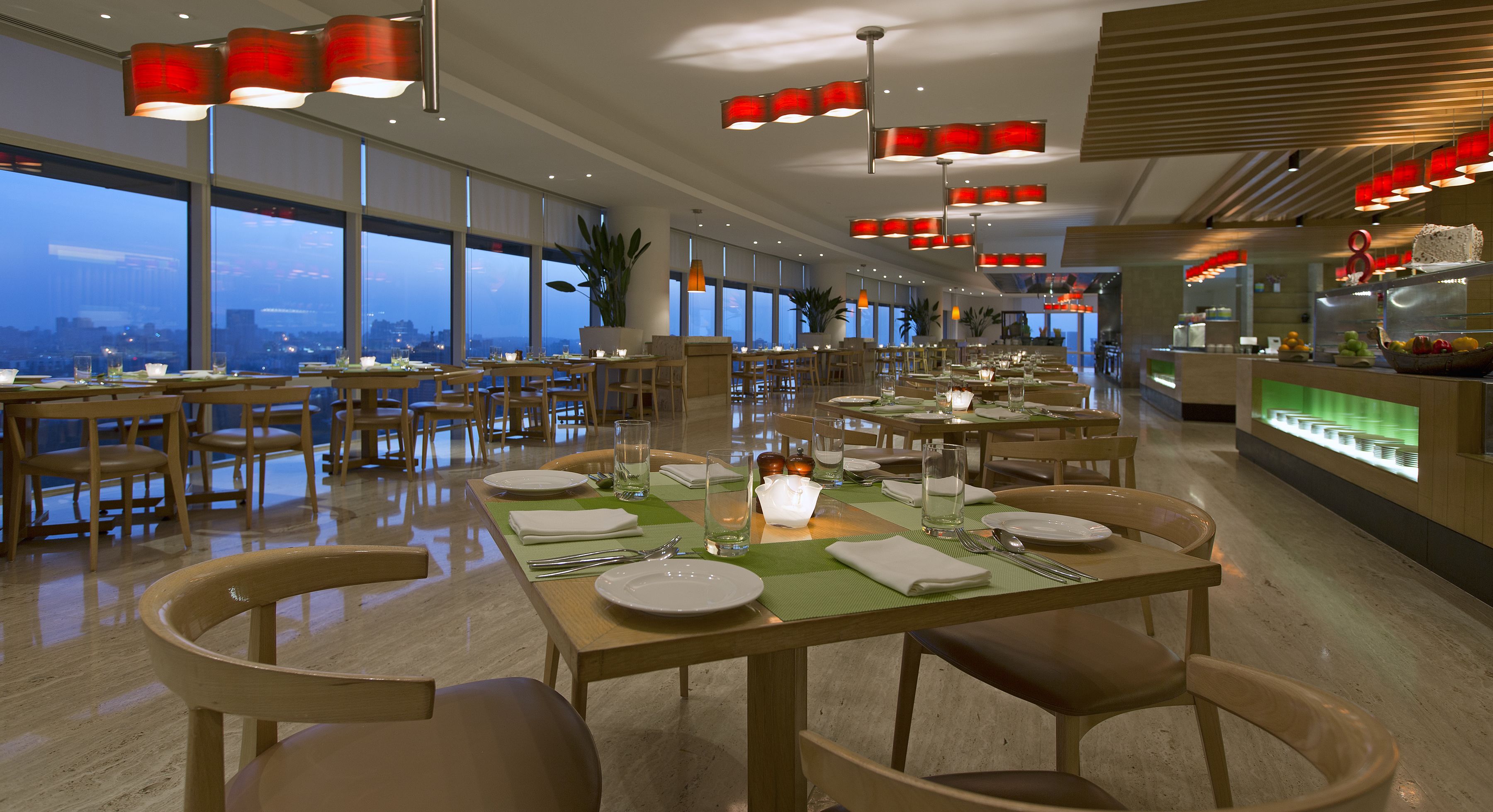 A restaurant with multiple dining tables set for guests, large windows offering a city view, and modern red pendant lighting.