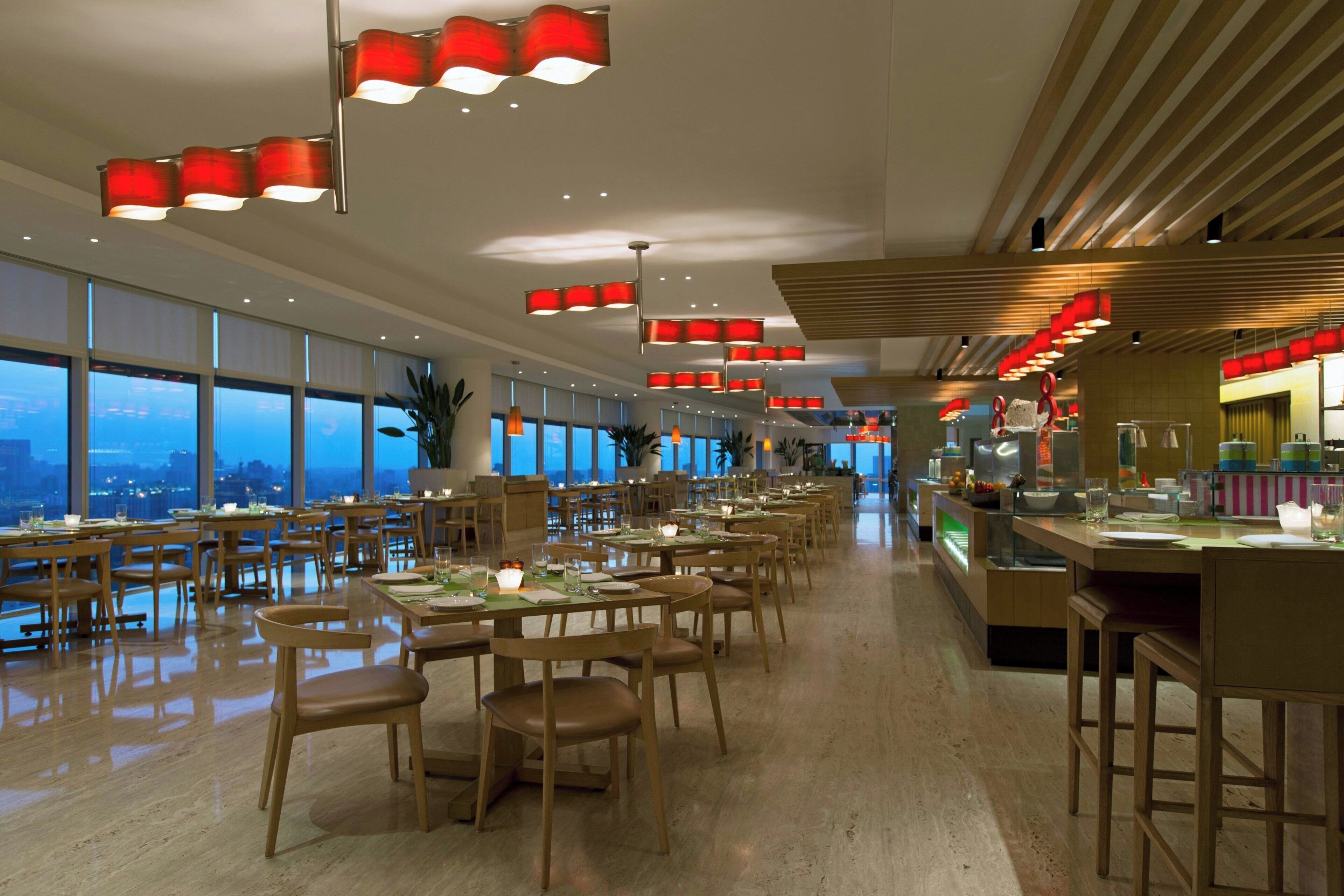 A spacious restaurant with multiple dining tables and chairs, large windows offering a view, and modern red pendant lighting.