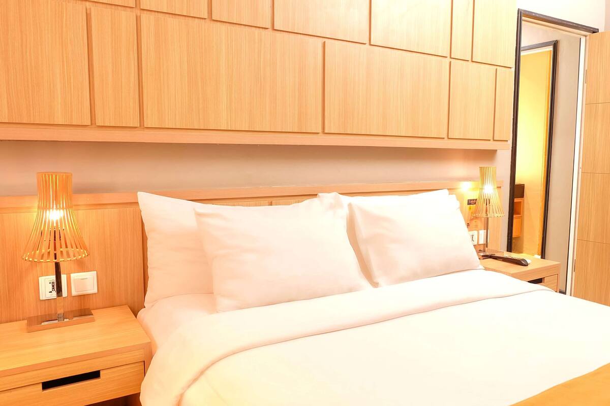 A hotel room with a wooden headboard, a bed with white bedding, a nightstand with a lamp, and a wardrobe.