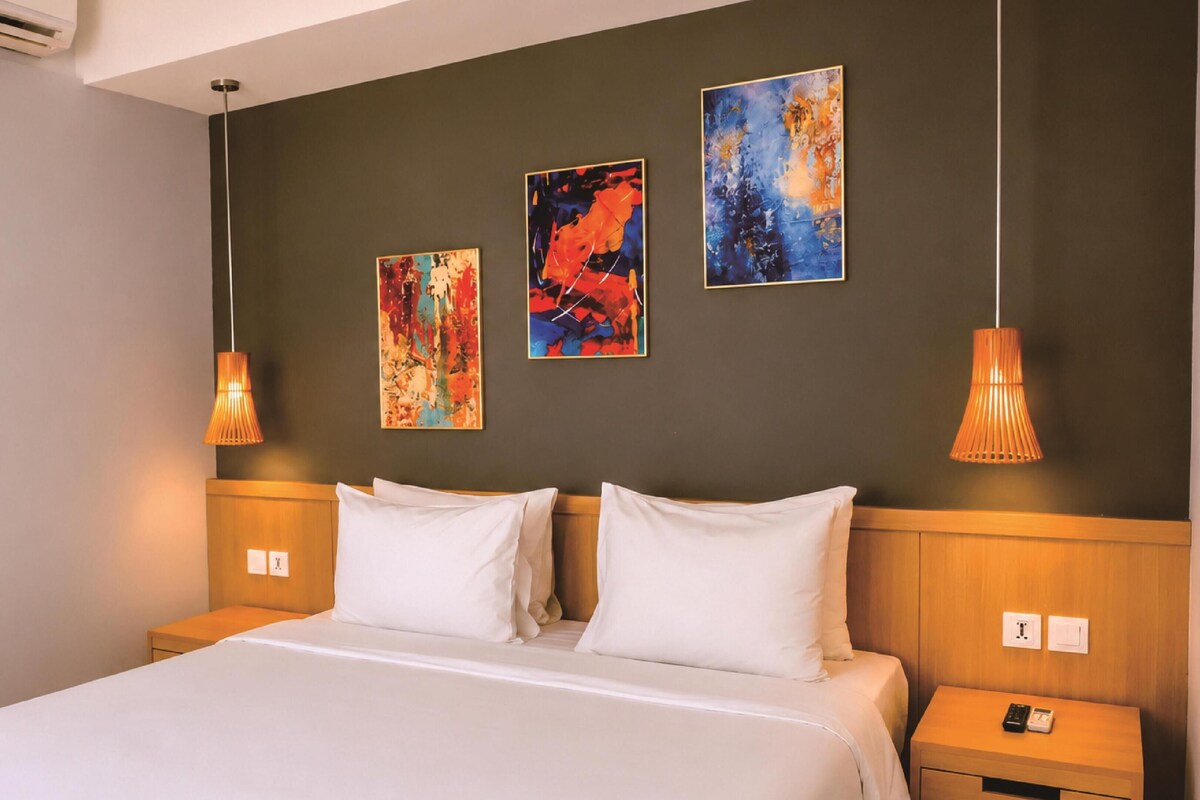 A hotel room with a bed, two bedside tables, a wall-mounted air conditioner, and three abstract paintings.