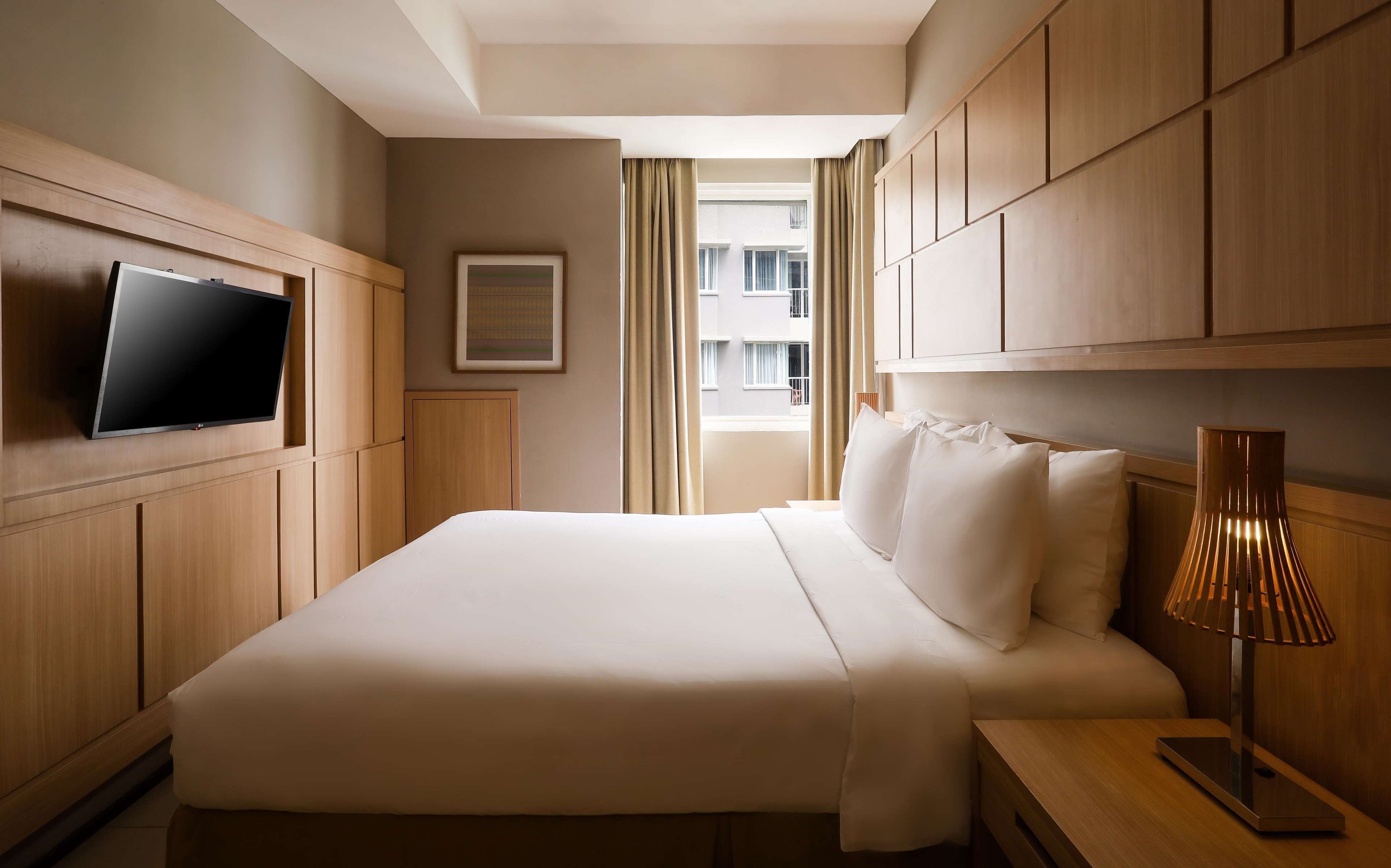 Une chambre d’hôtel avec un lit, un téléviseur fixé au mur, un tableau encadré et une fenêtre avec des rideaux.