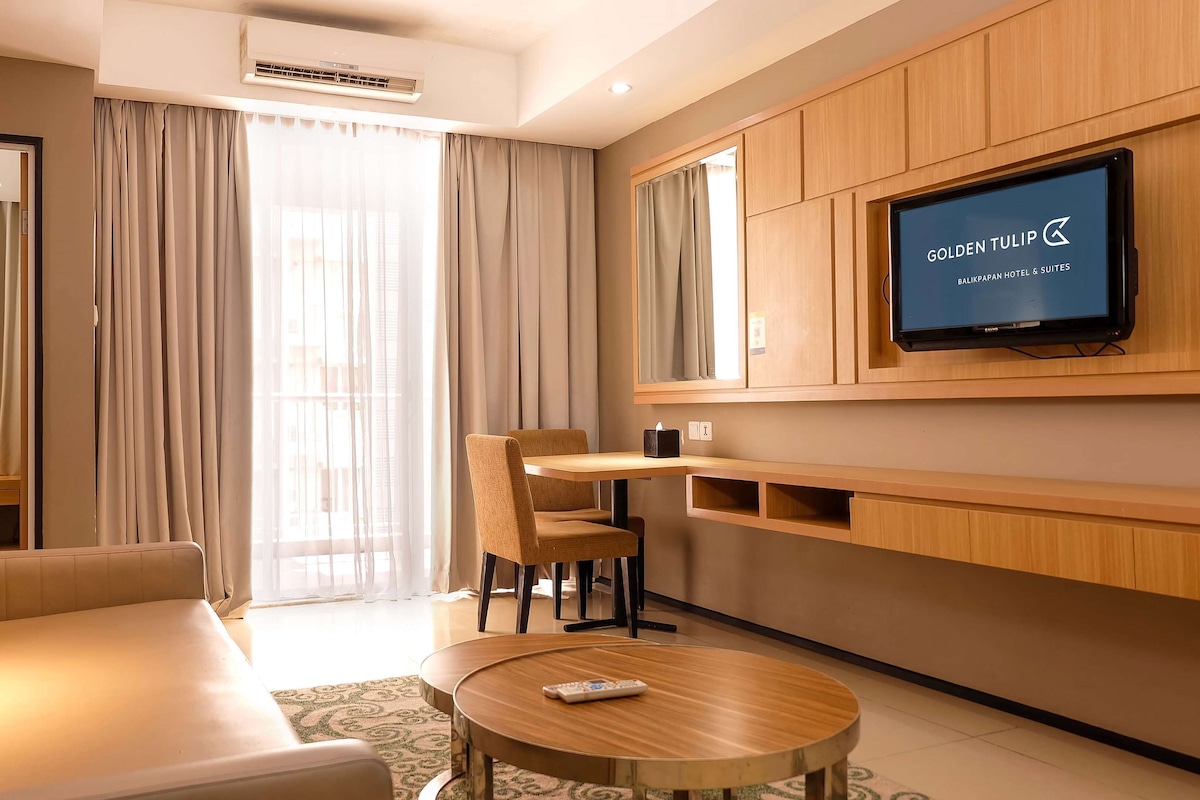 A modern hotel room with a sofa, a dining table with chairs, a TV mounted on the wall, and a large window with curtains.