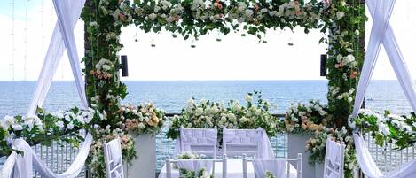 An outdoor wedding setup with white chairs and a floral arch, overlooking the sea.