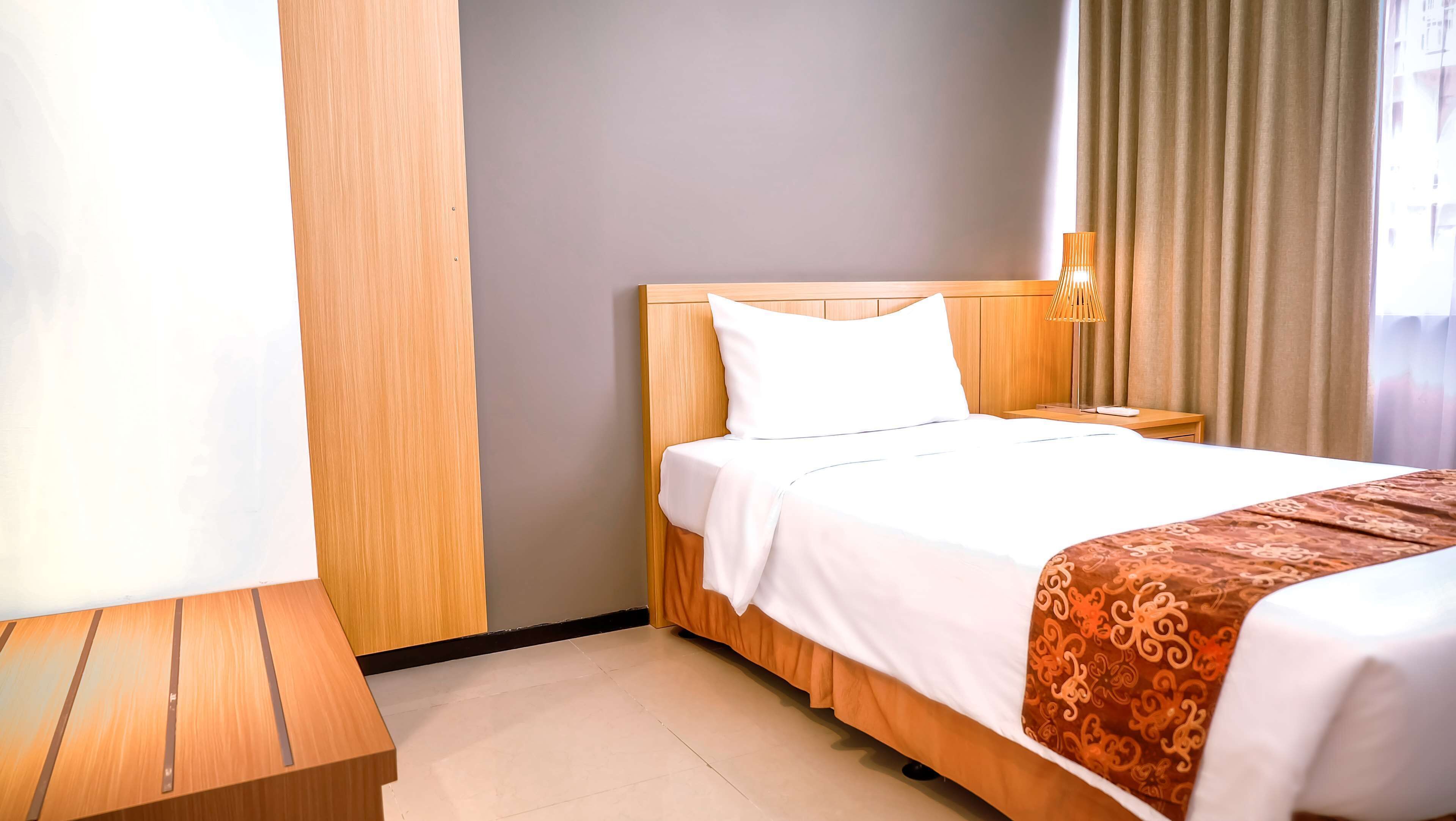 Une chambre d’hôtel avec une tête de lit en bois, un lit avec une literie blanche et marron, une table de chevet avec une lampe, et une vue sur l’extérieur par une fenêtre avec des rideaux.