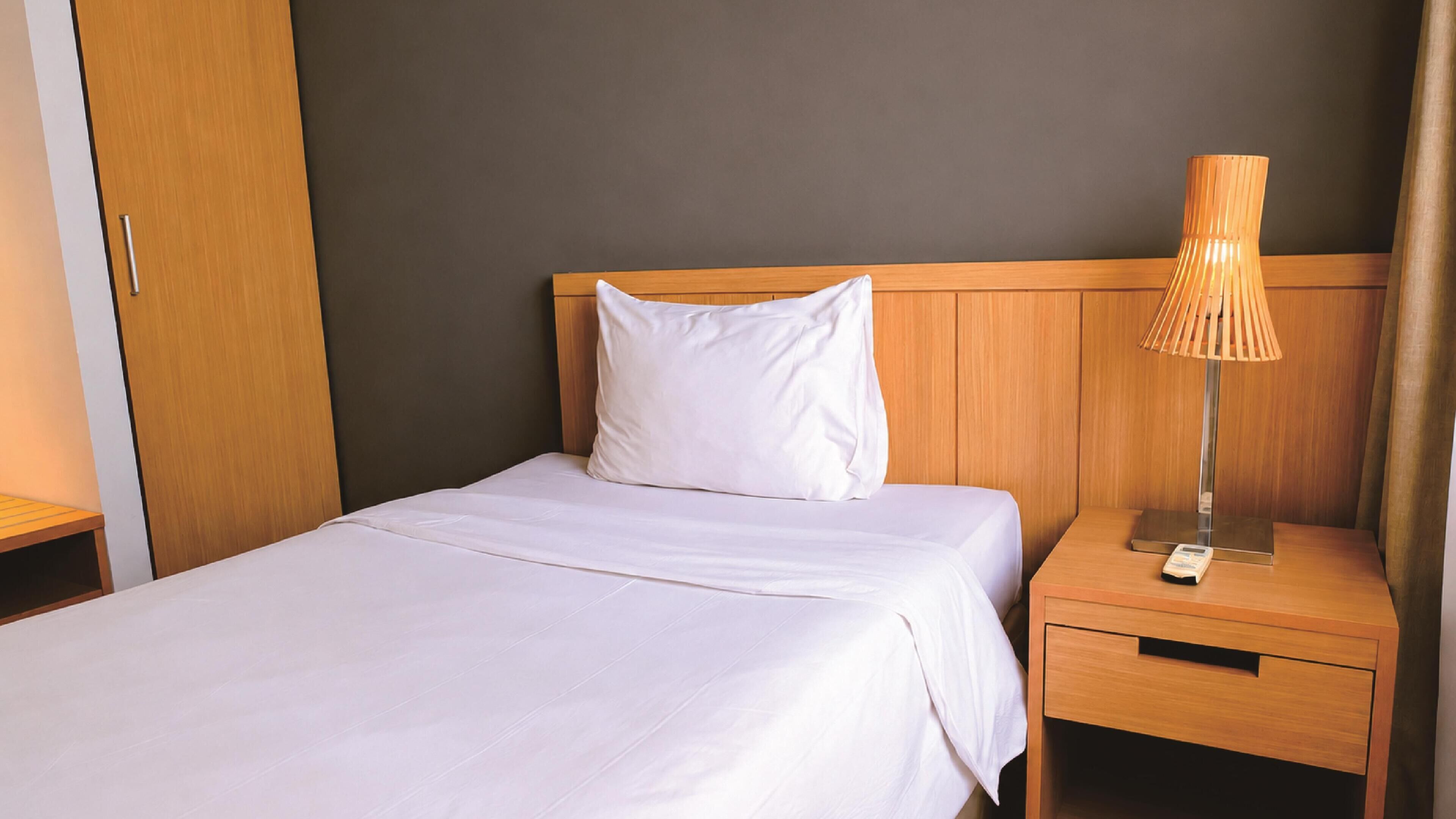 Una cama bien hecha con sábanas blancas, cabecera de madera, lámpara de noche y mesita de noche con cajón.