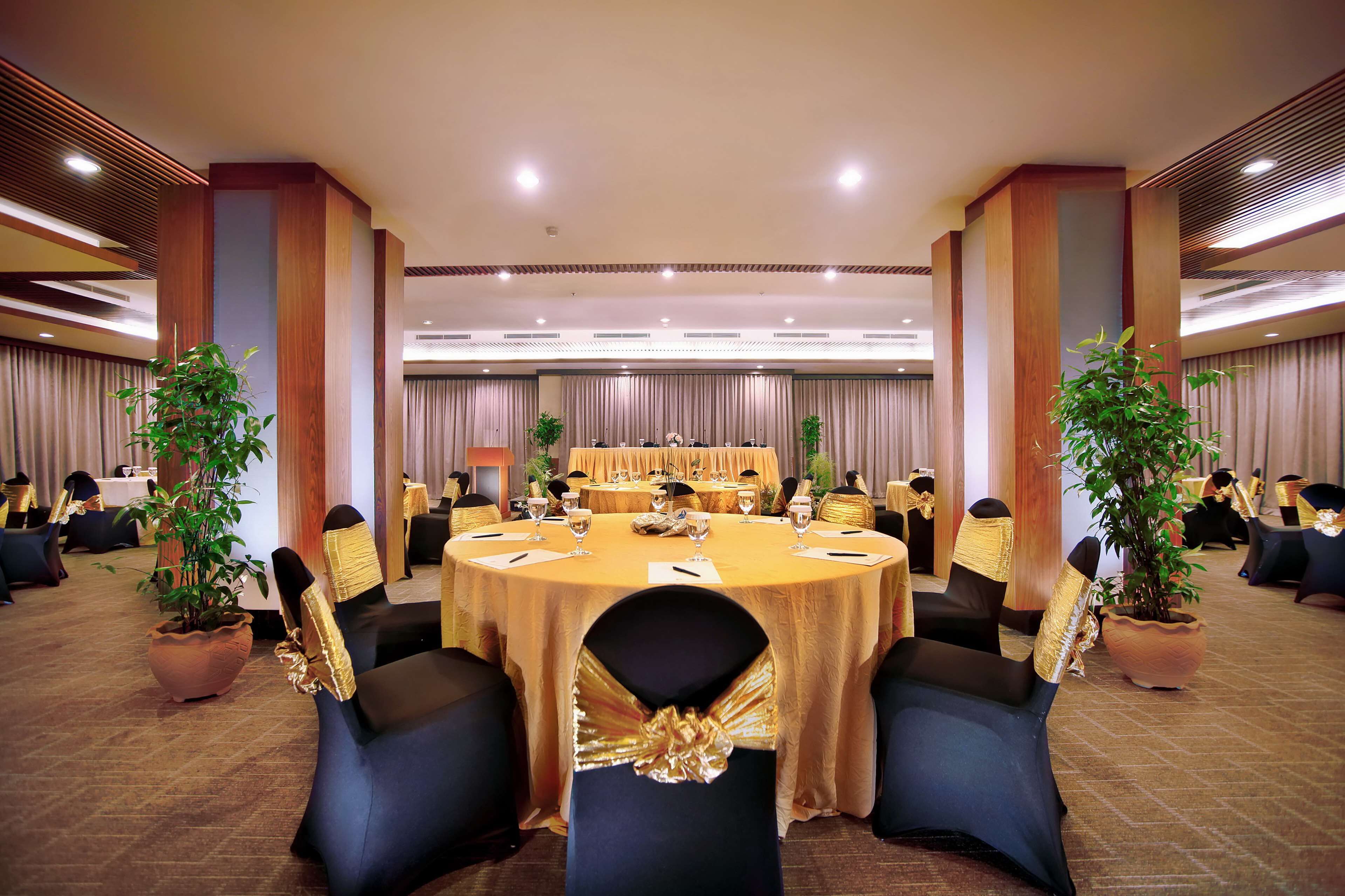 Un salón de conferencias con mesas redondas, sillas negras con cintas doradas y plantas en macetas.