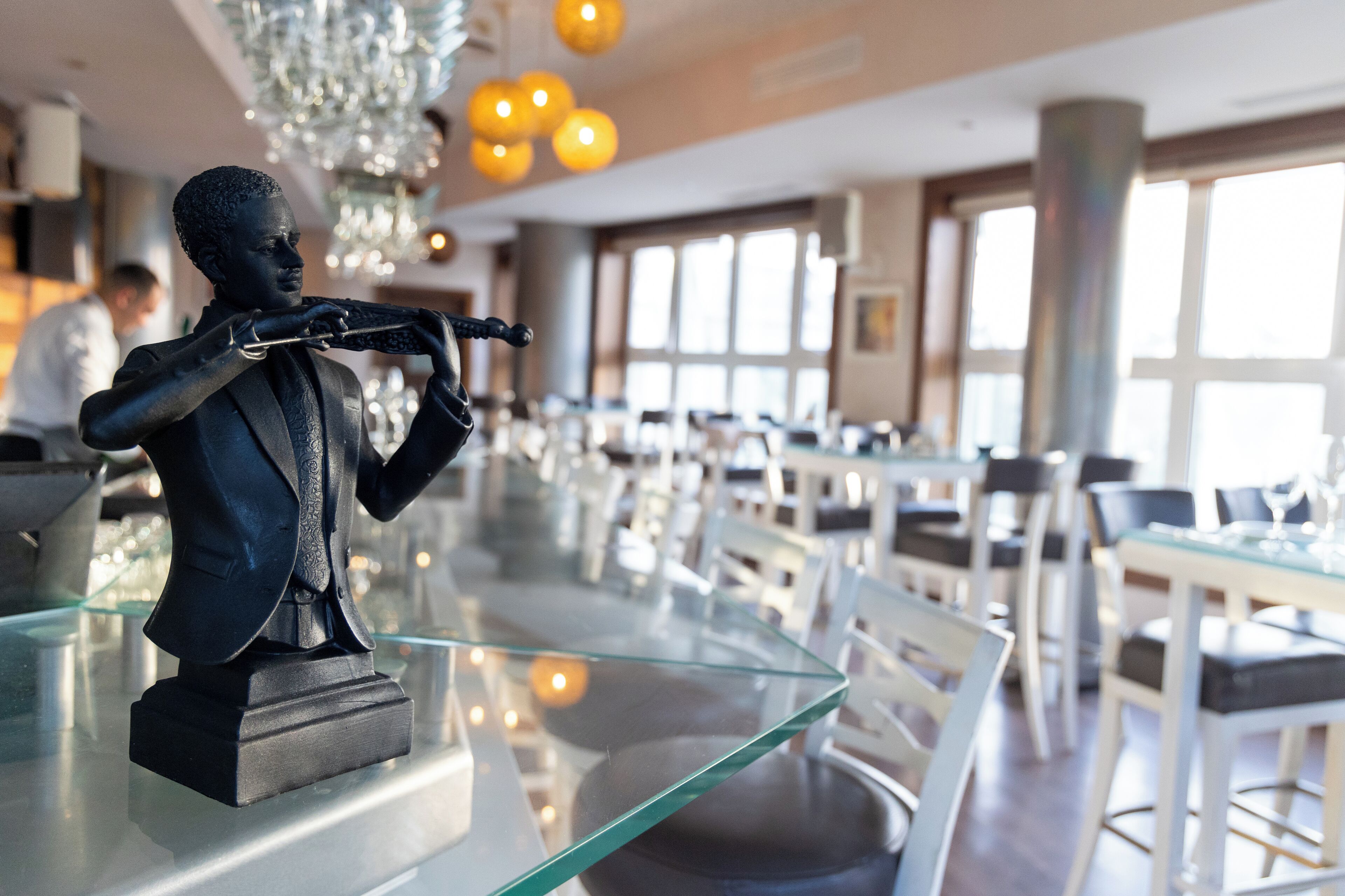 A statue of a man playing a trumpet on a glass table in a modern restaurant with white chairs and large windows.