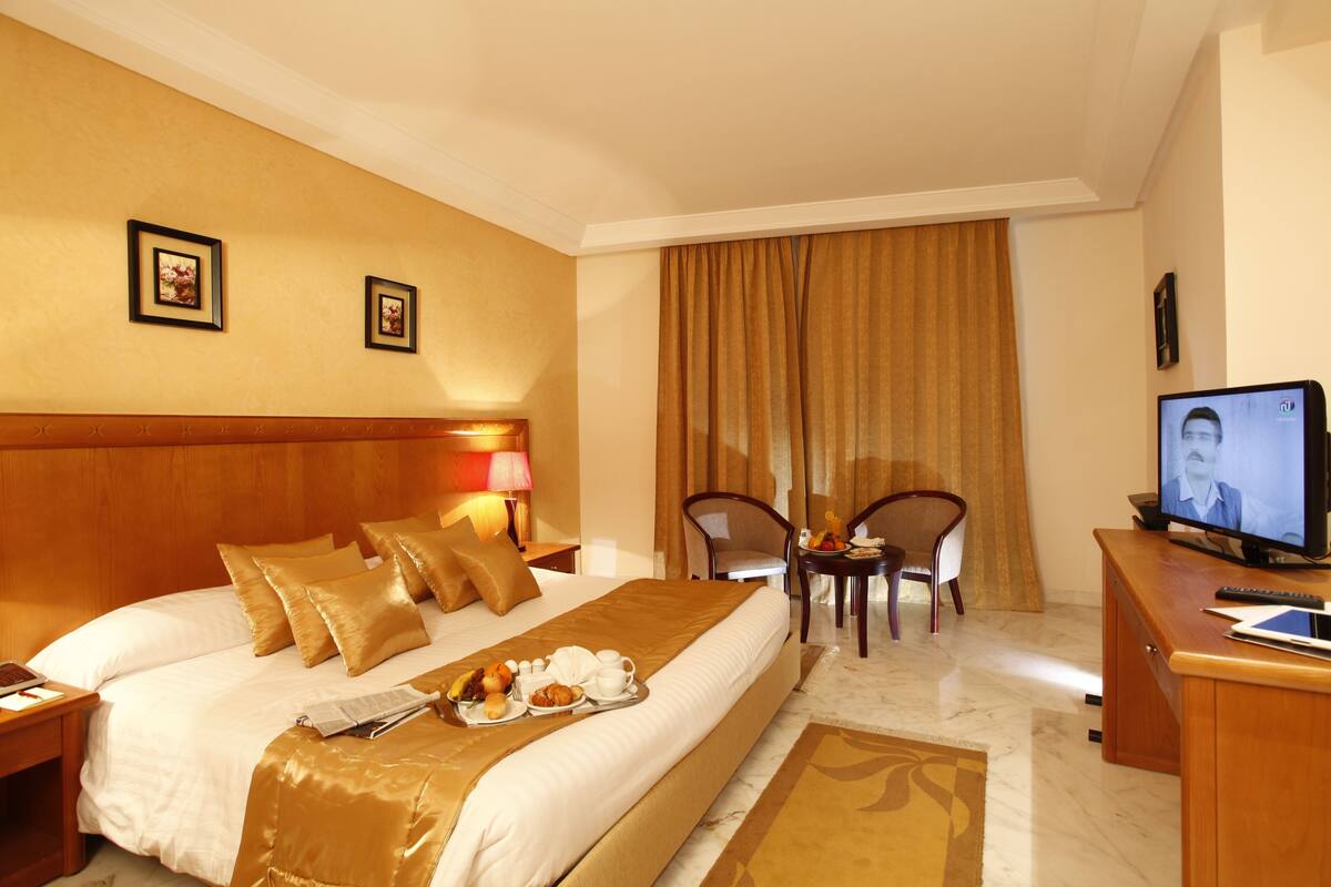 A hotel room with a large bed, a desk with a computer, a television, and a dining area with chairs and a table.