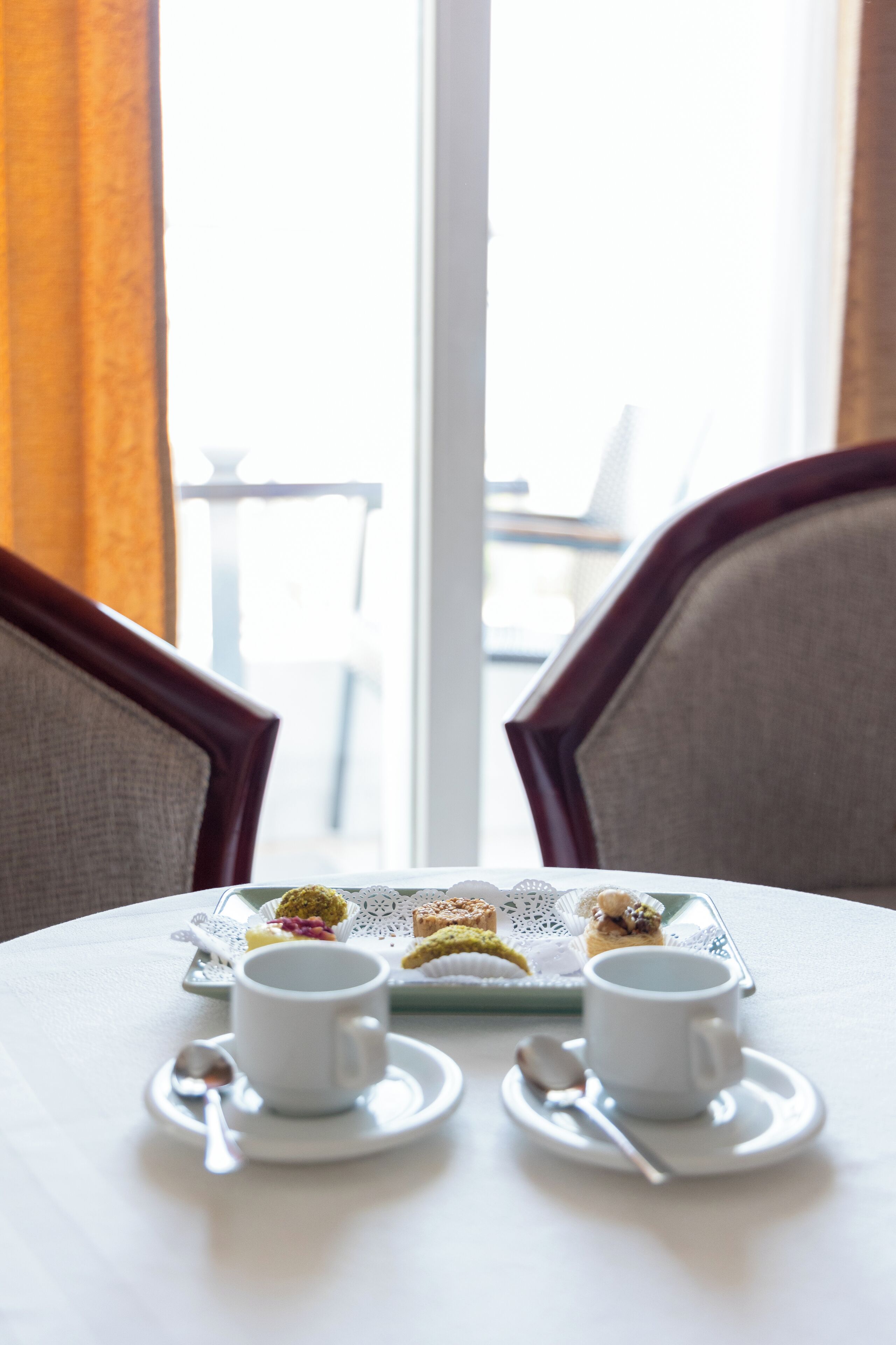 Una mesa con dos tazas blancas y una bandeja de pasteles, frente a una ventana con cortinas.