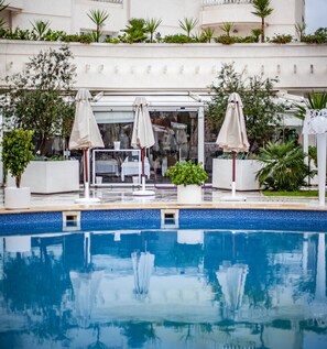 A swimming pool with lounge chairs and umbrellas, surrounded by greenery and a building facade.