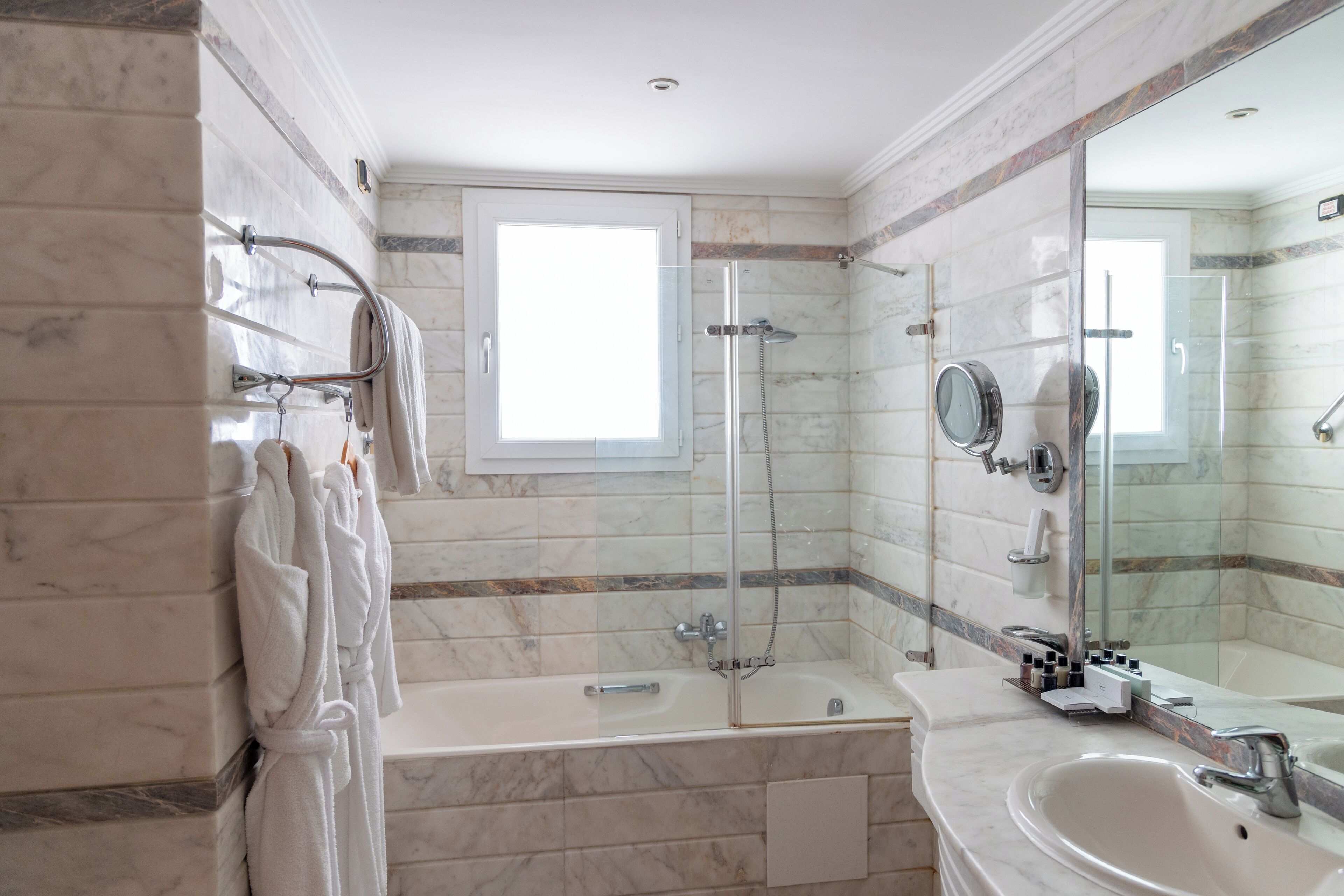 A modern bathroom with a glass-enclosed shower, a bathtub, a large mirror, and marble walls.