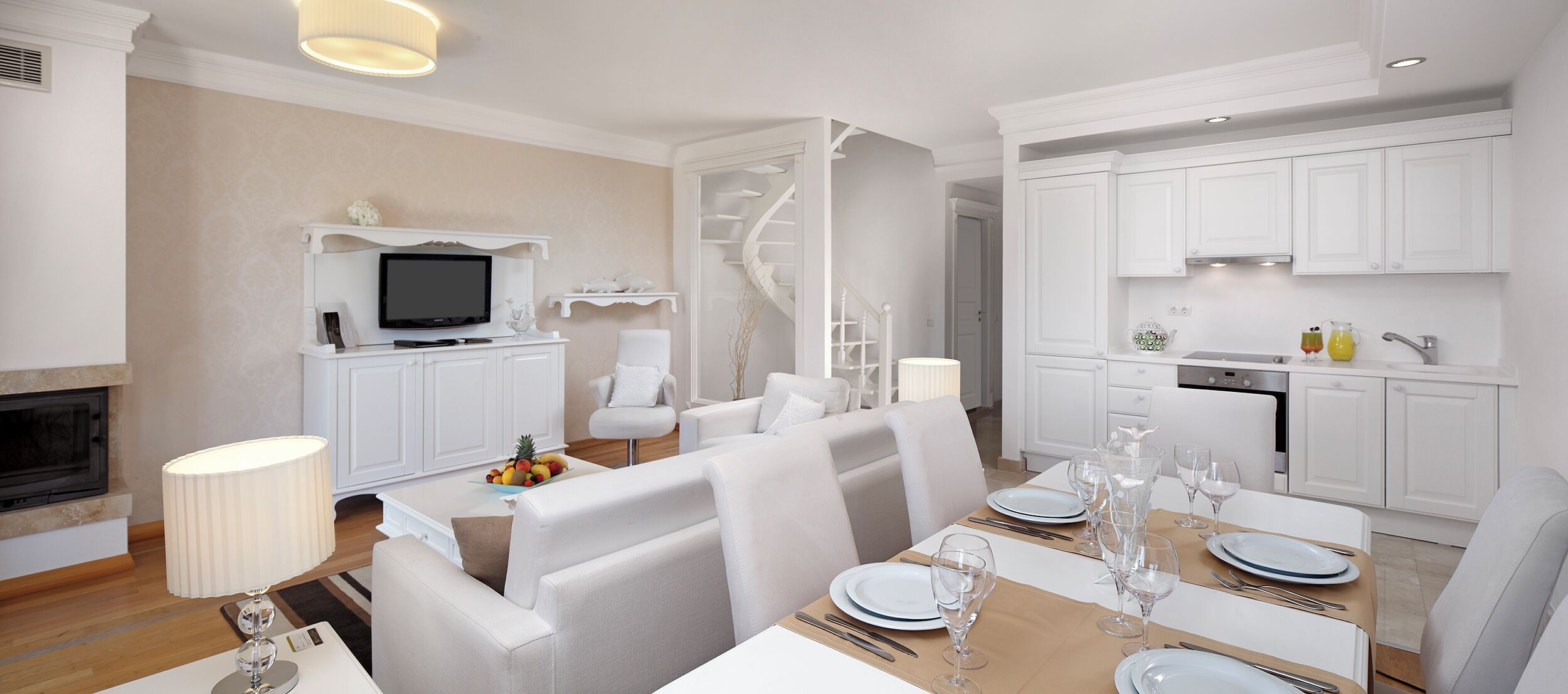 A modern, well-lit dining area with a wooden table set for four, white upholstered chairs, and a television mounted on a white cabinet.