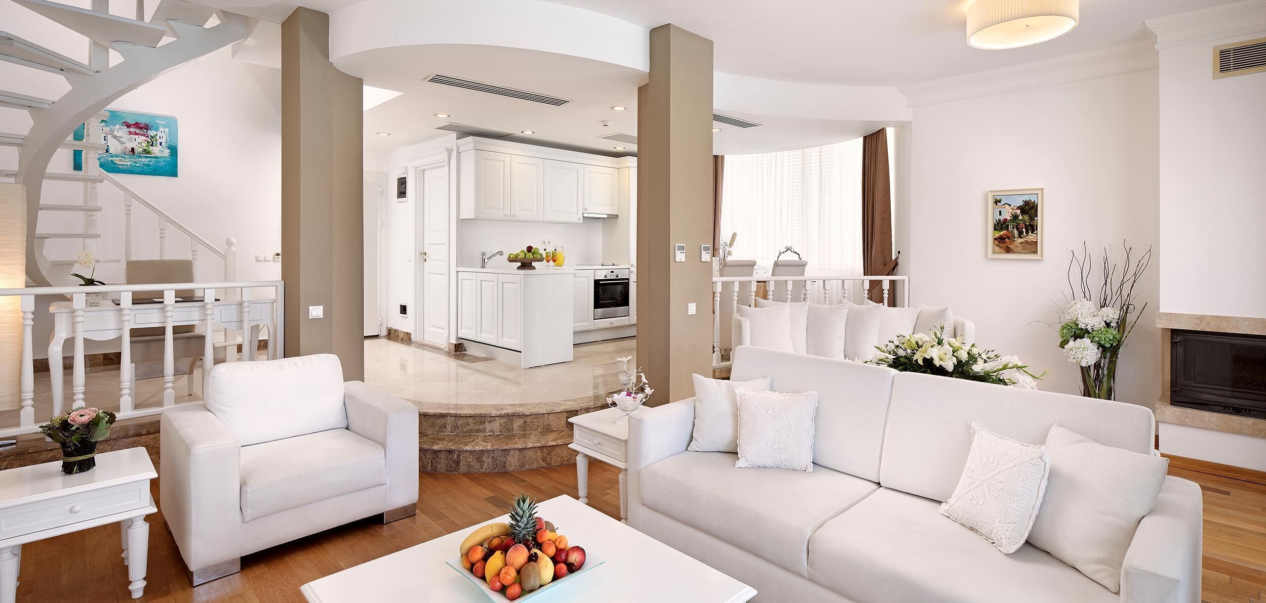 A modern living room with white furniture, a fruit bowl on a coffee table, and a staircase leading to a dining area.
