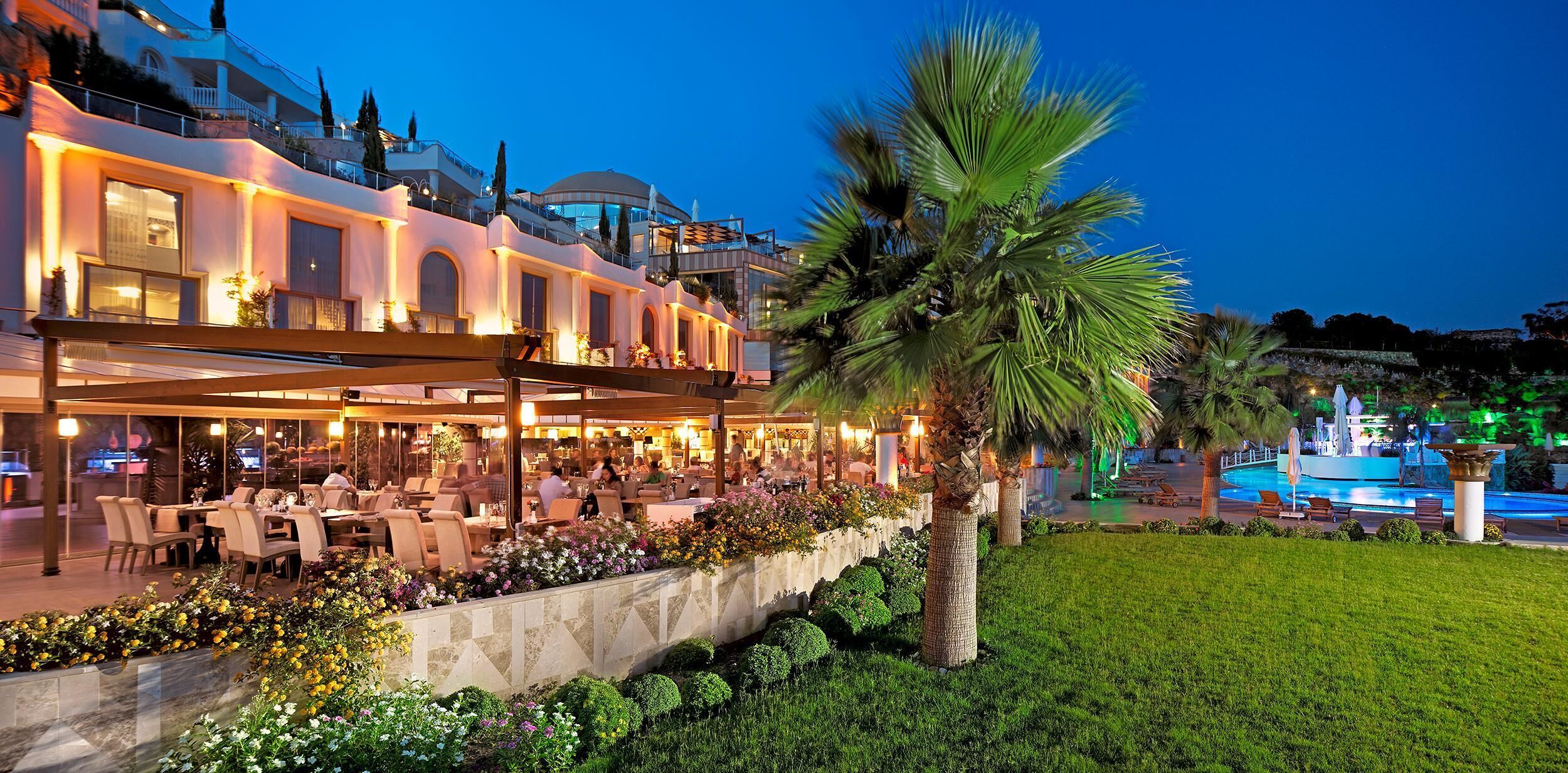 Un área de comedor al aire libre bien iluminada, con mesas y sillas, rodeada por un jardín ajardinado con palmeras y flores.