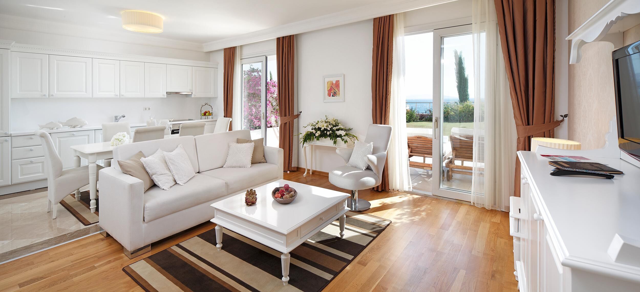 Uma sala de estar moderna com um sofá branco, uma mesa de centro e uma área de jantar com uma mesa e cadeiras brancas. Há uma varanda com vista para o mar.