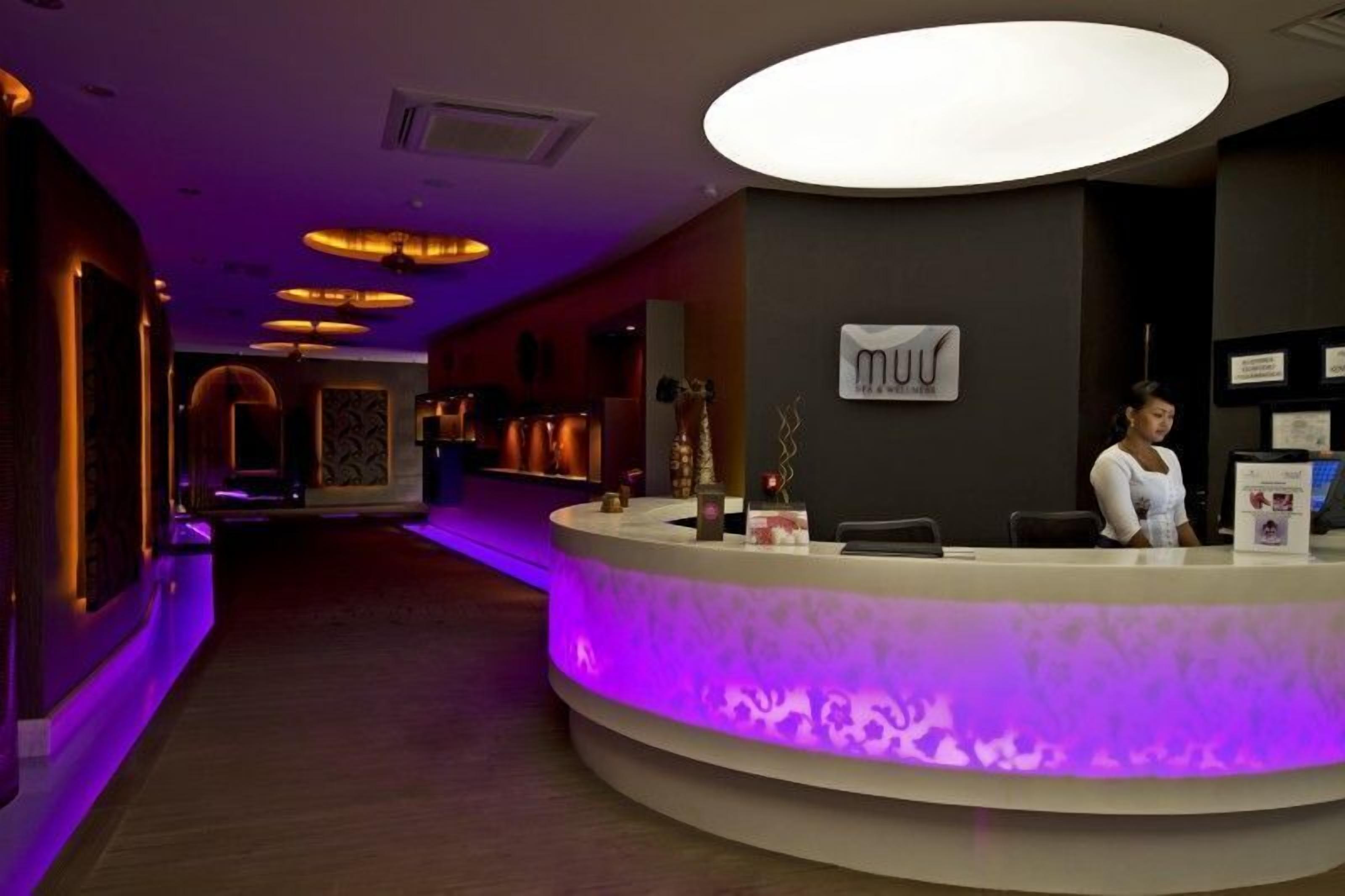 En hotellobby med modern design, med en receptionsdisk vars fot är upplyst i lila, en stor, upplyst skylt ovanför disken och en hall med trägolv och dekorativ belysning.