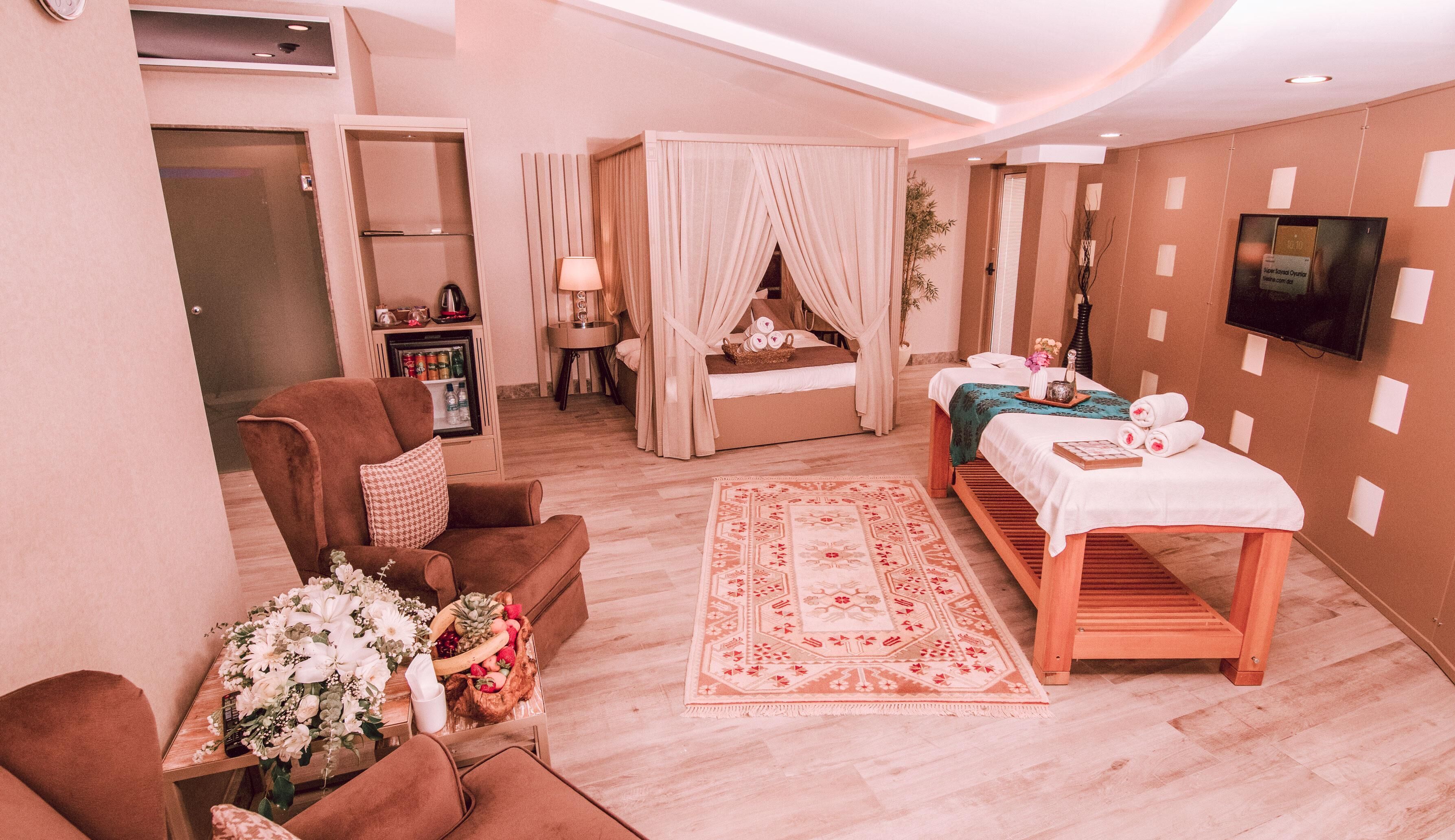 A hotel room with a bed, a massage table, a sofa, a small table with flowers, a TV, and a minibar.